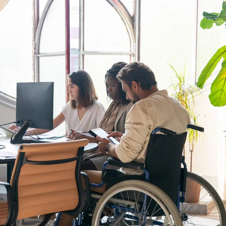 Three diverse colleagues, working together at a table with a computer and a tablet in a bright office.