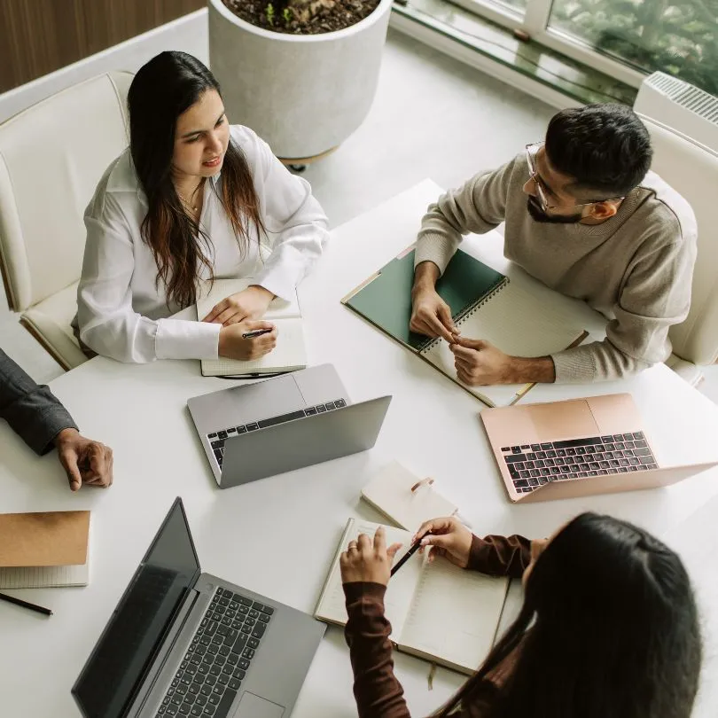 Four professional people sitting around a white table with laptops and notebooks, engaged in a discussion.