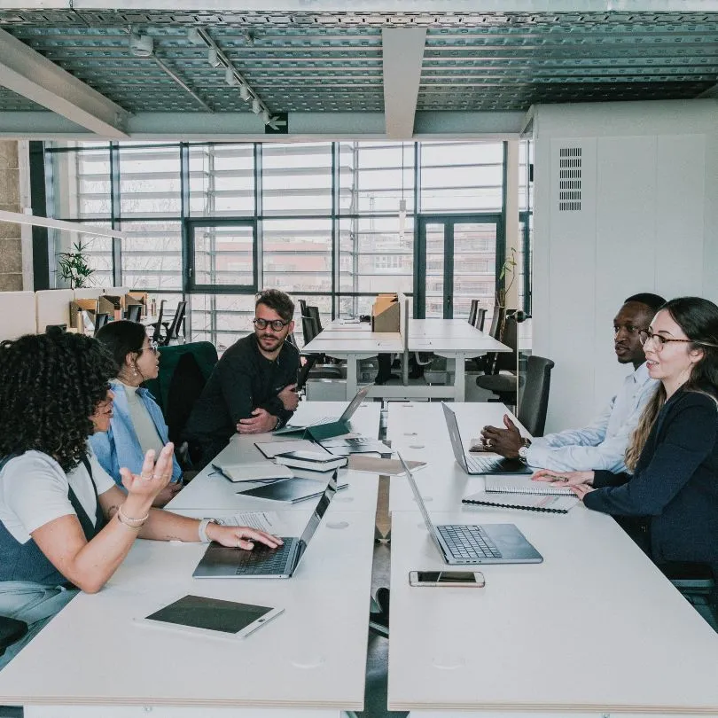 Five professional colleagues engaged in a meeting at a modern office, sitting across a long white table with laptops and notebooks.