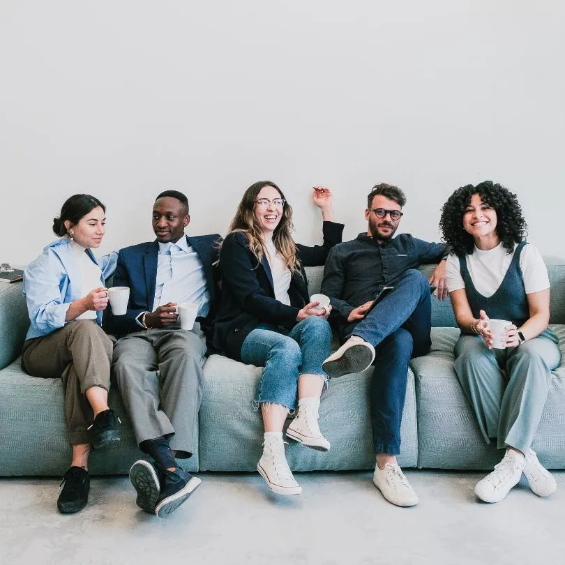 Five diverse young professionals sitting on a gray couch, smiling and holding white coffee mugs.