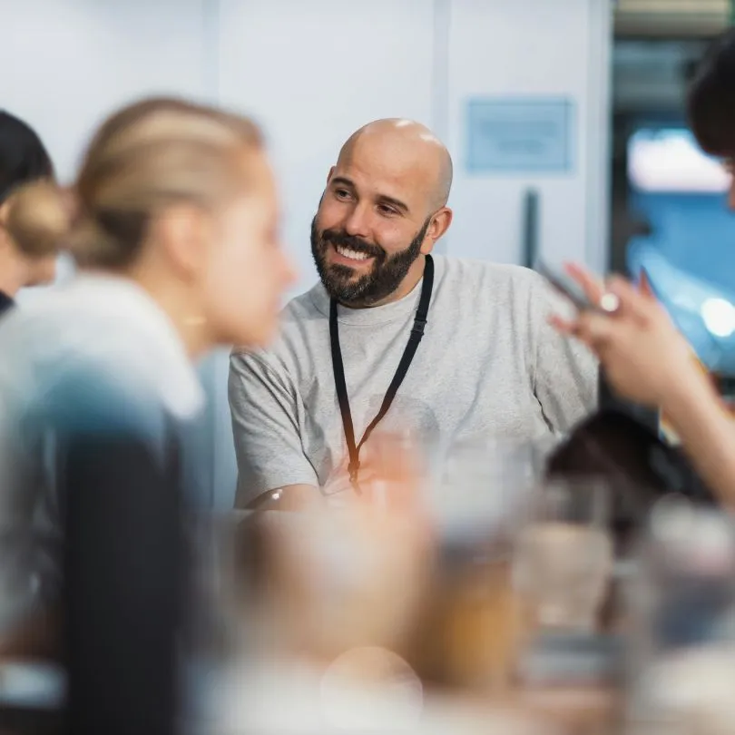 Smiling business man with a beard wearing a gray shirt and black lanyard, sitting at a table in a casual indoor setting.