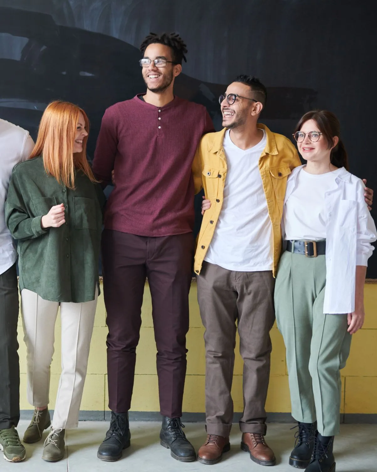 Group of four diverse young adults standing side by side, smiling and embracing each other in casual clothing.