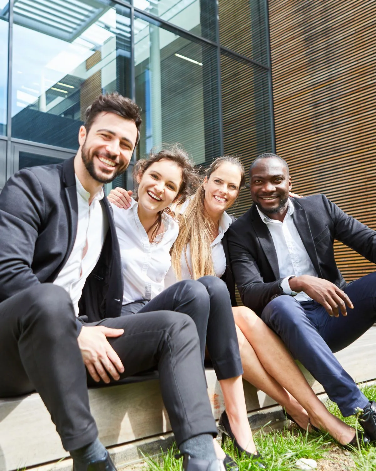 Four diverse young professionals sitting outside smiling and posing for a group photo.