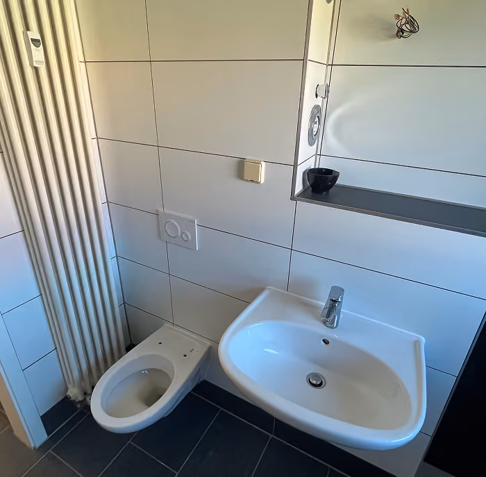 Kleines Badezimmer mit weißer Toilette und Wandwaschbecken, großen Fliesen und Heizkörper.