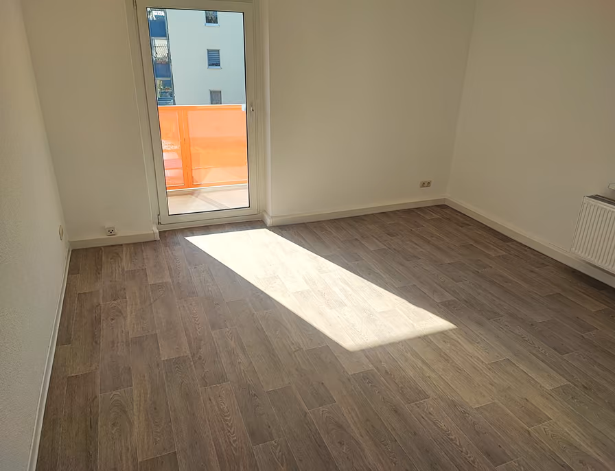 Leerer Raum mit Holzfußboden, weißen Wänden und einer Tür zu einem Balkon mit orangefarbenem Geländer.
