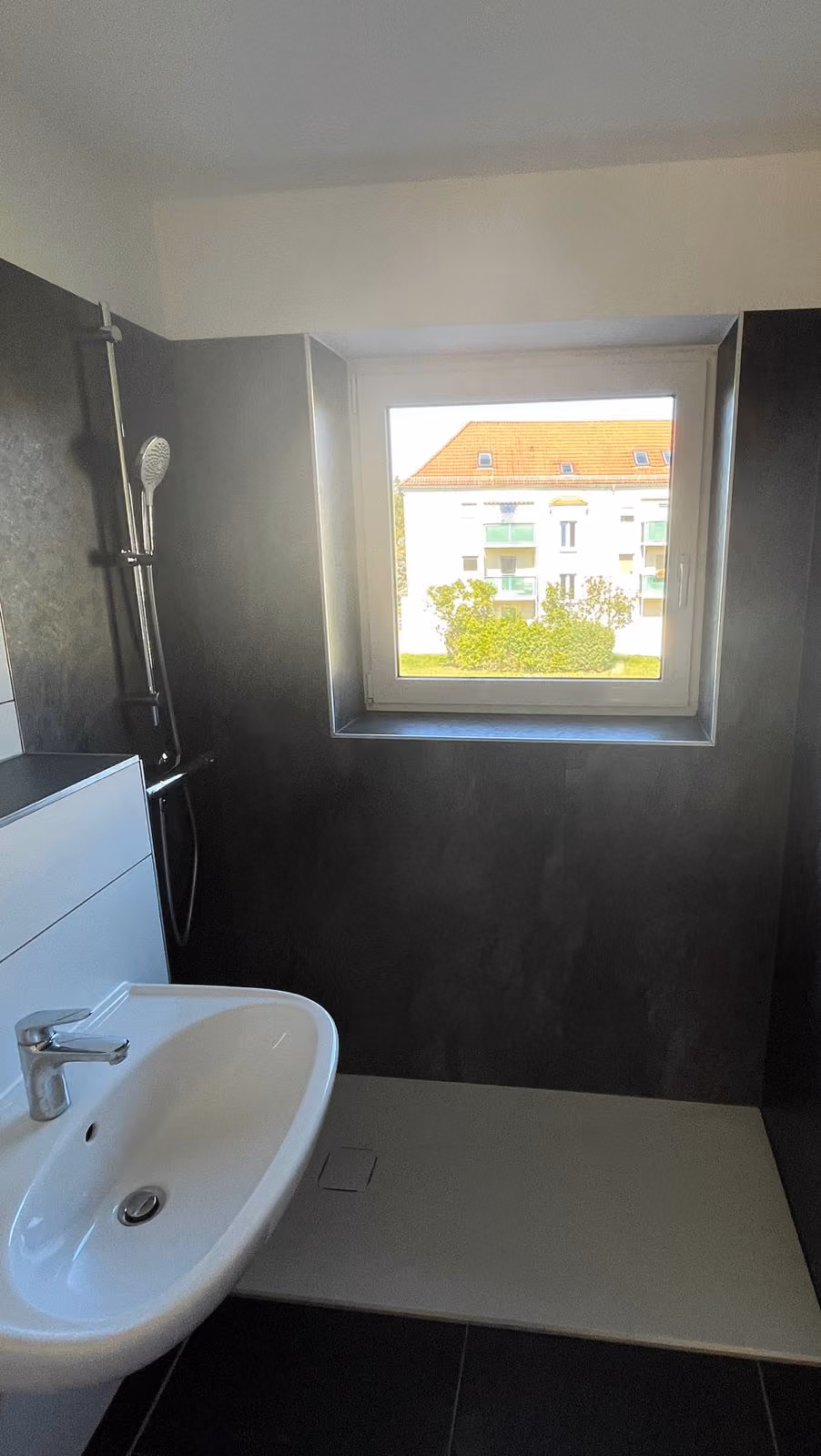 Modern bathroom with white sink, black walls, showerhead, and window showing a building outside.