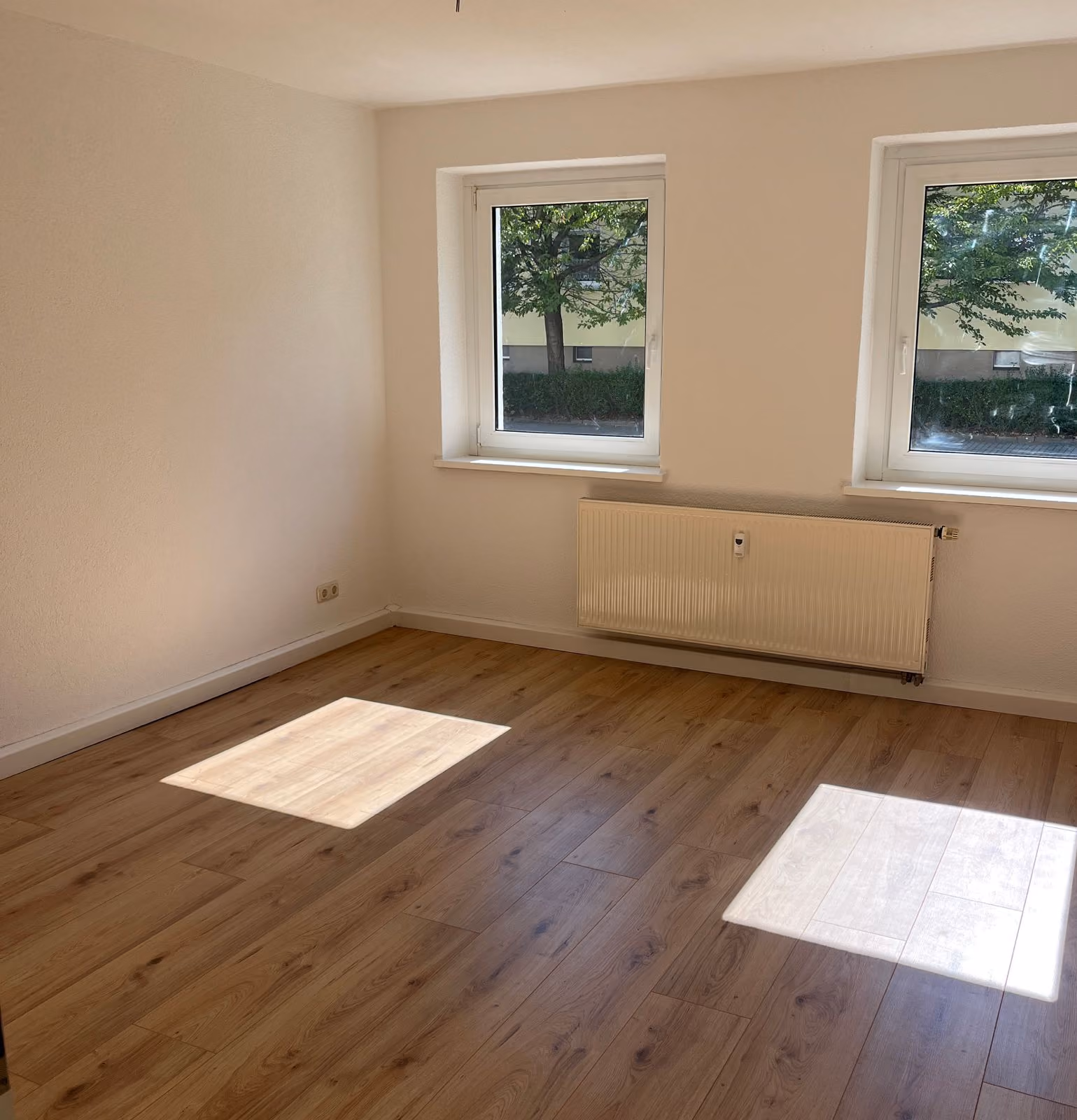 Leerer Raum mit hellen Holzboden, zwei Fenstern und Heizung an der Wand.
