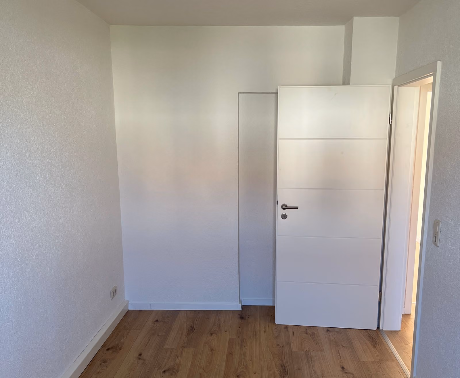 Leerer Raum mit weißen Wänden, Holzboden und einer offenen weißen Tür.