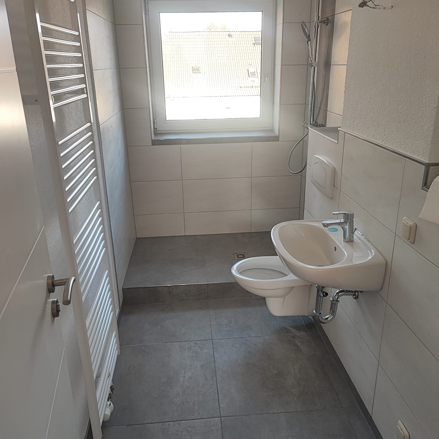 Kleine moderne Badezimmer mit grauen Fliesen, Waschtisch, WC, Dusche und Fenster.