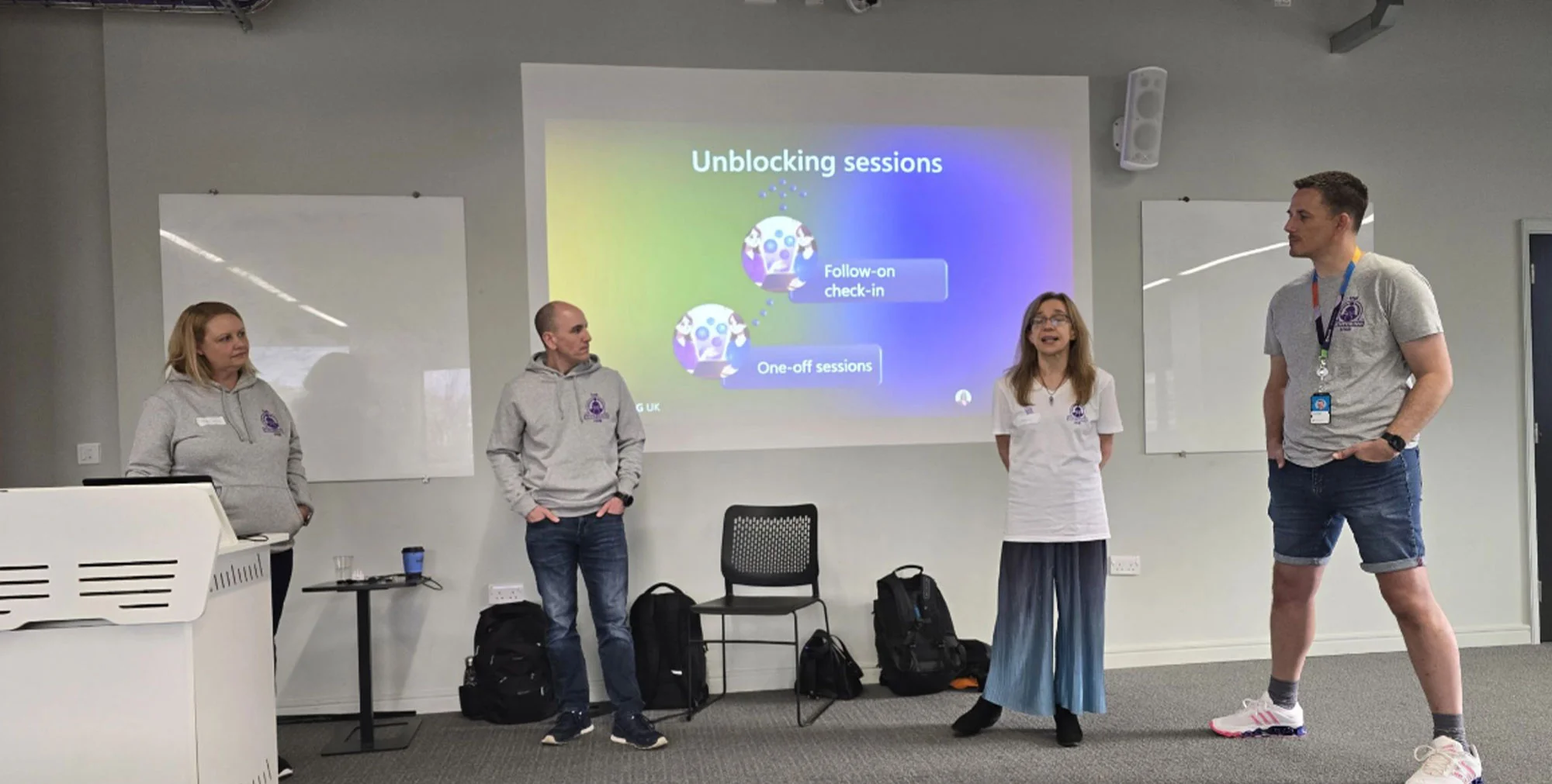 Two women and two women presenting in front of a projector screen, displaying a diagram about unblocking sessions.