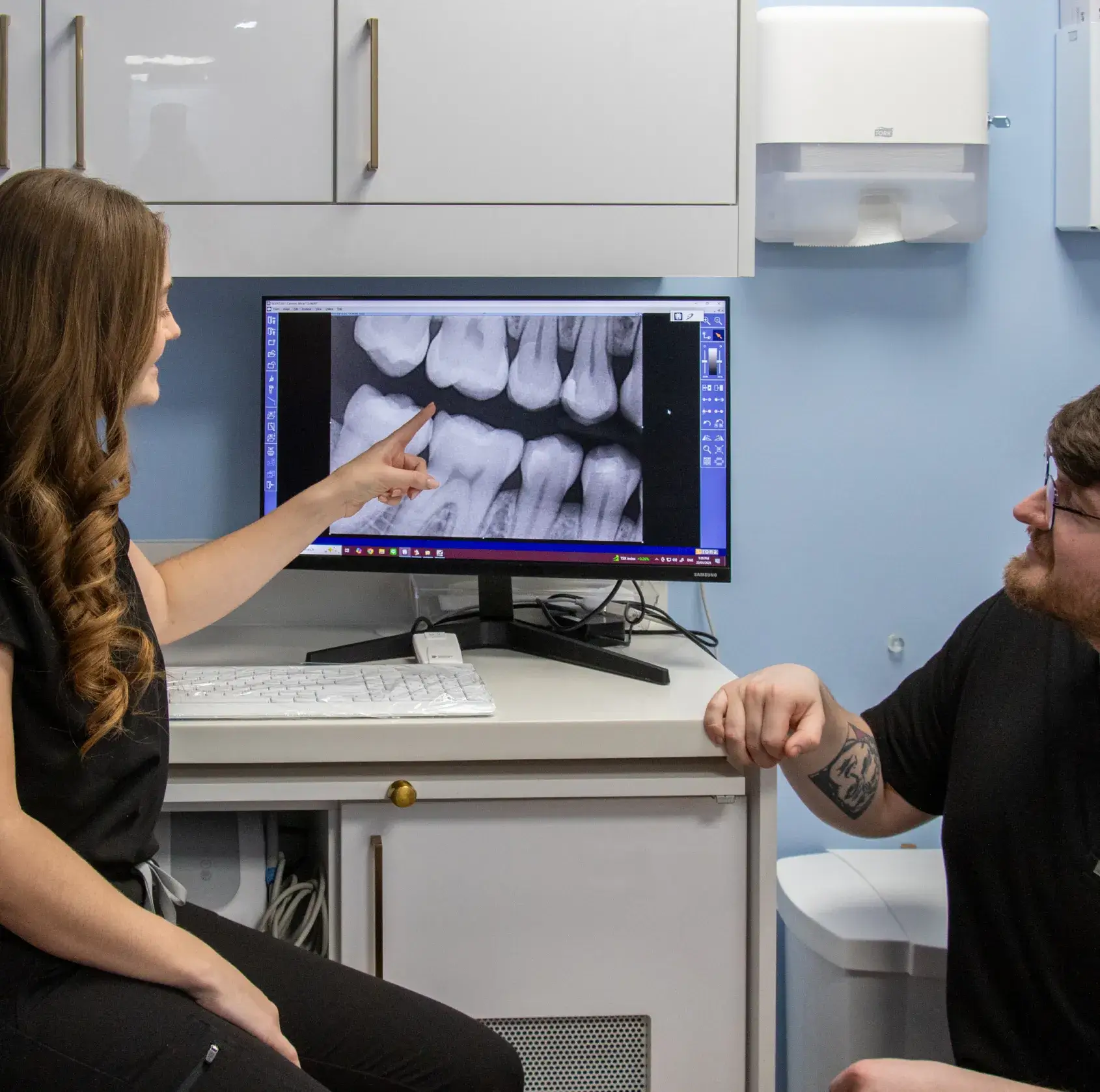 A dental professional shows a patient an X-ray image of teeth on a computer screen.