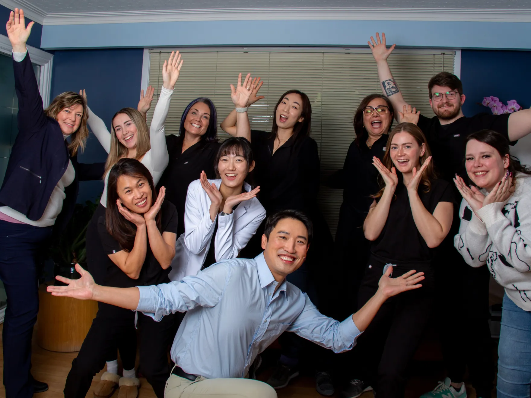 Group of diverse people smiling and posing cheerfully with raised arms and hands near faces in an indoor setting.