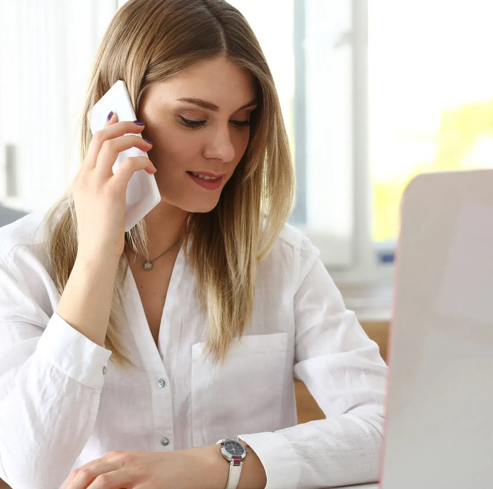 A woman in a white shirt talks on her phone while sitting at a desk.