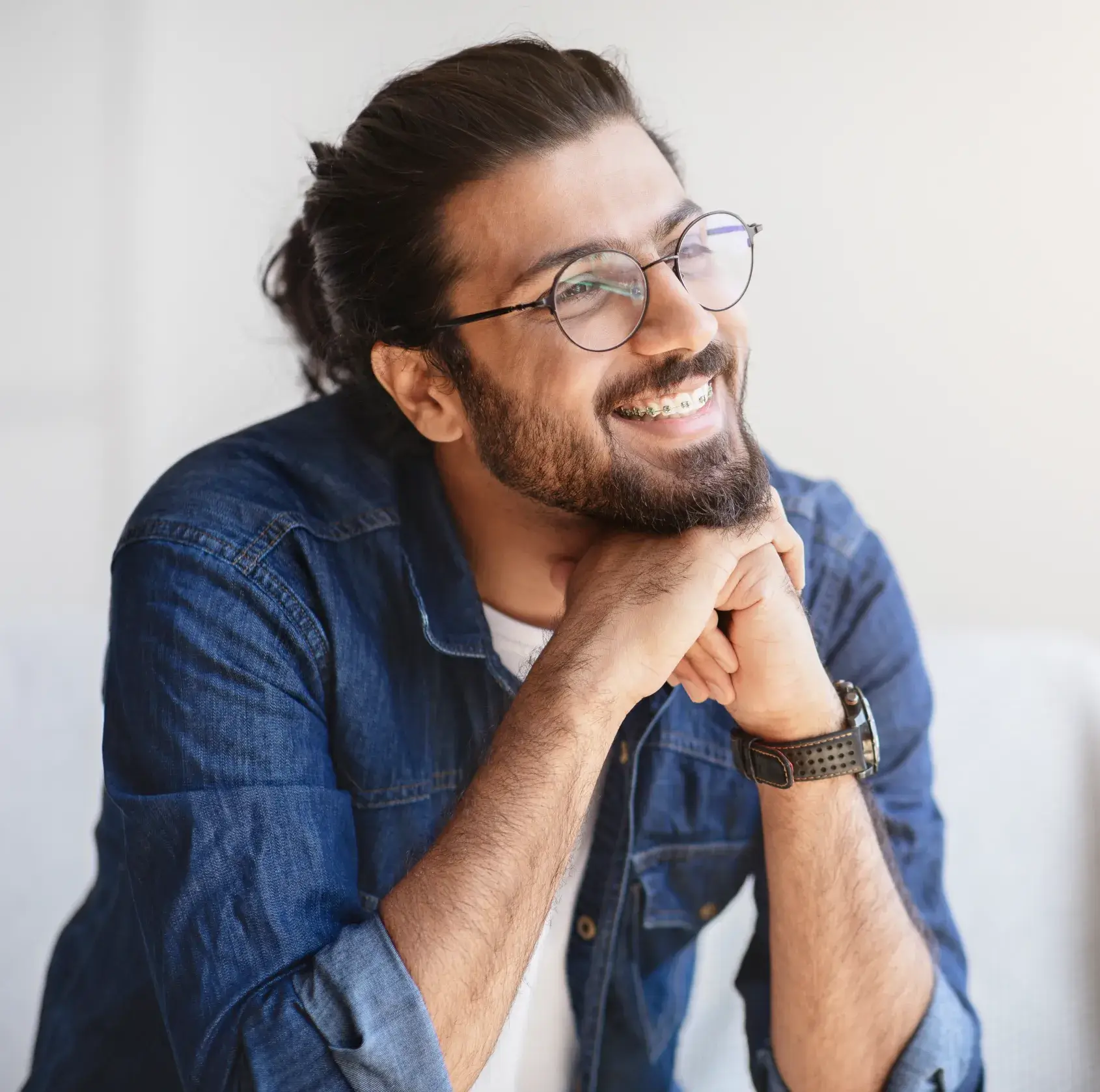 Man with glasses and braces, smiling, wearing a denim shirt and resting his chin on his hands.