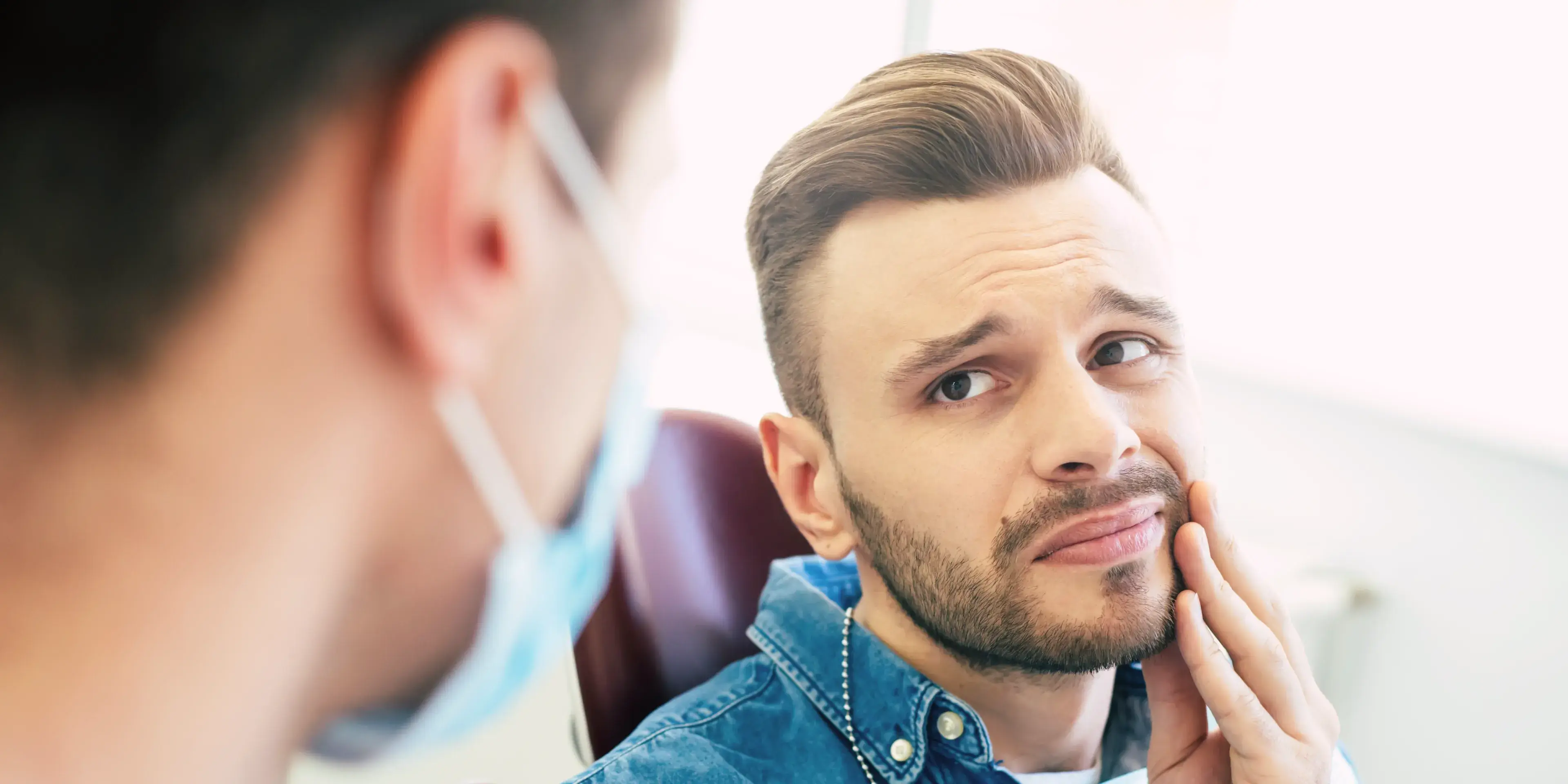 A man sits in a dental chair, looking concerned, and touching his cheek.