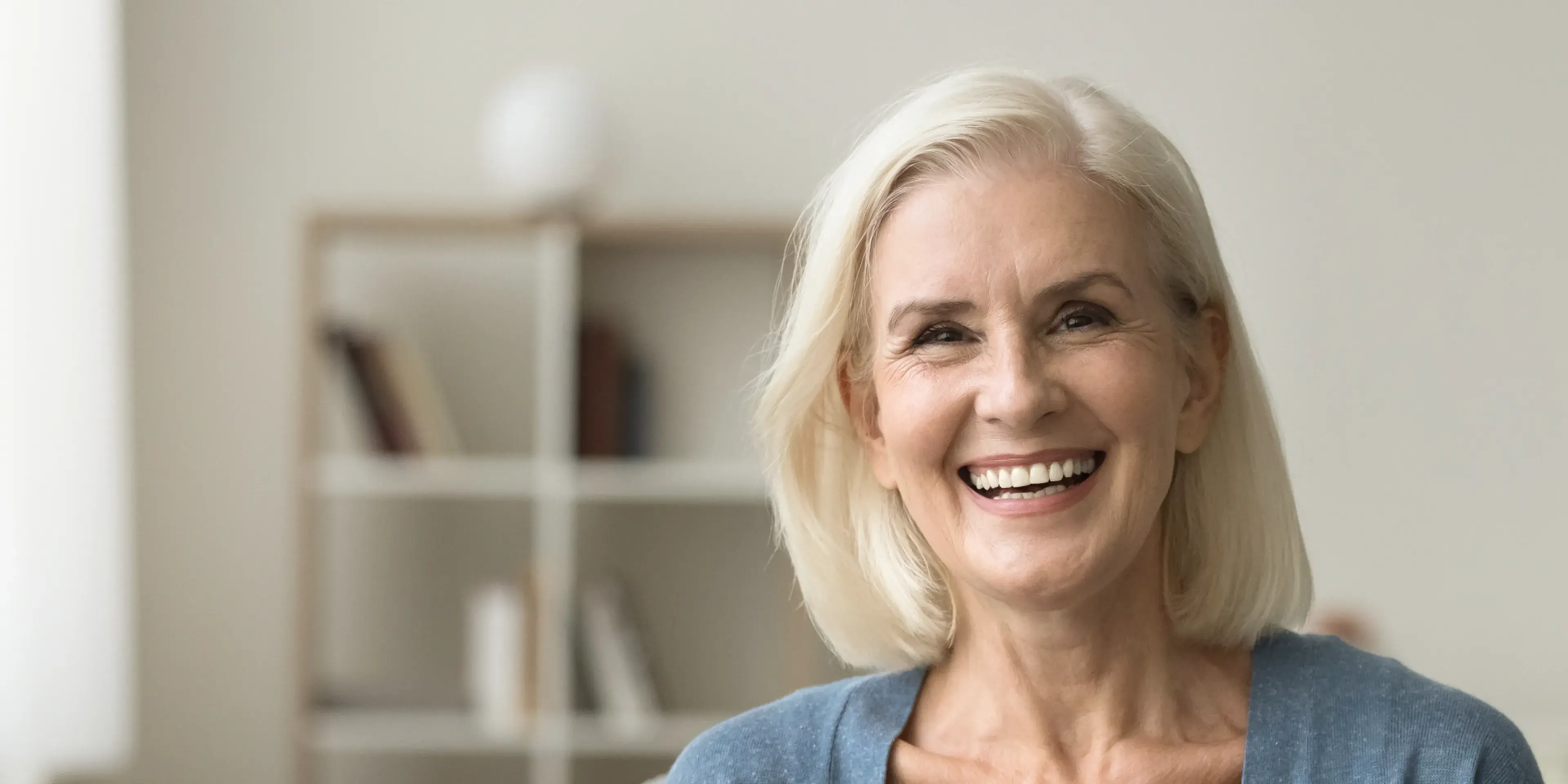 Smiling older woman with short white hair wearing a blue top in a room with shelves.