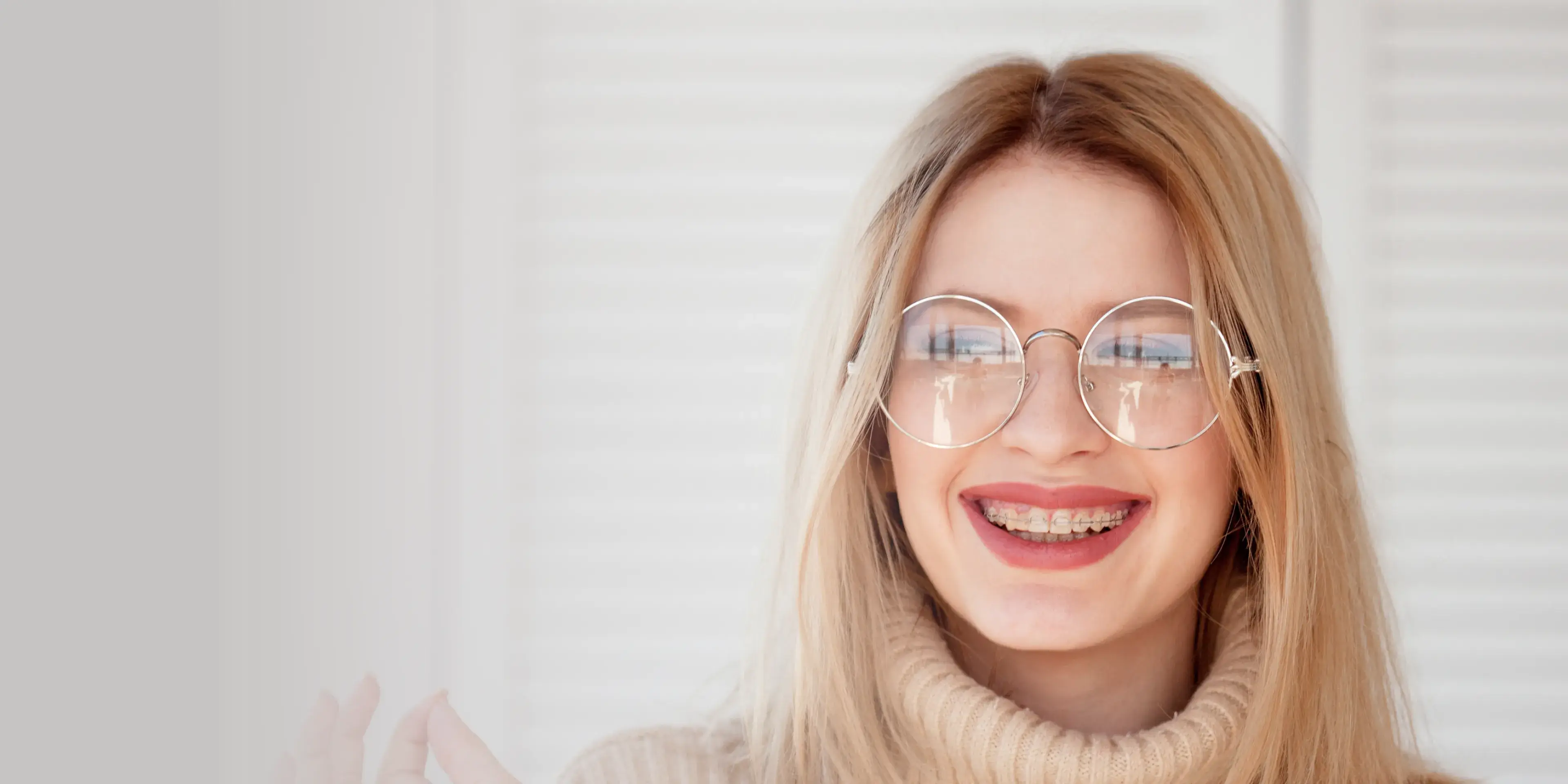 Smiling person wearing glasses and braces, dressed in a beige turtleneck sweater.