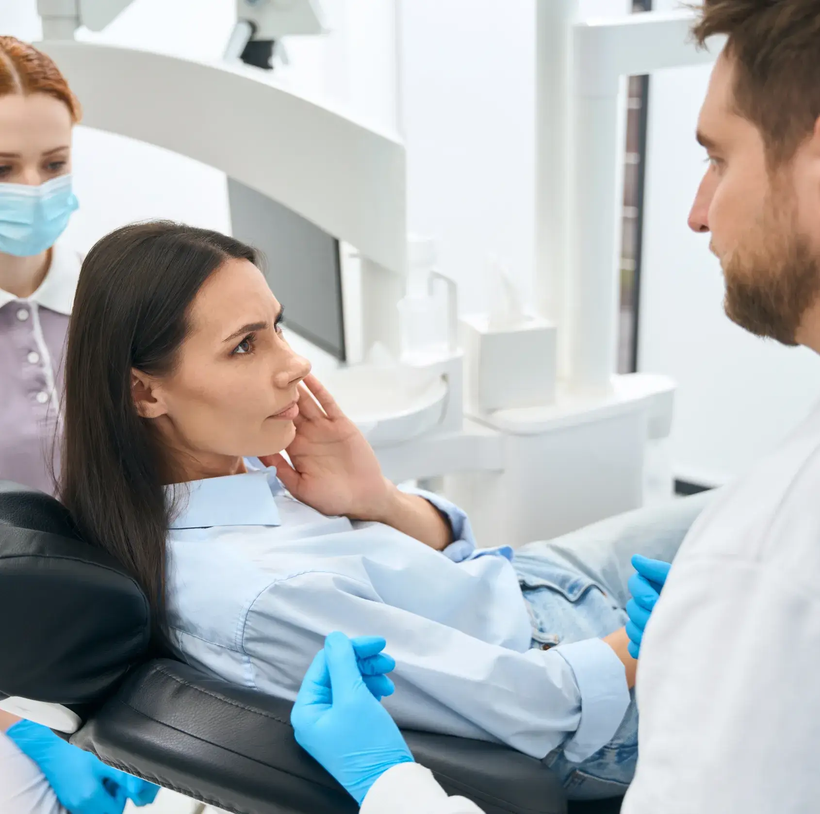 A woman sits in a dental chair, listening to a dentist and dental assistant discussing her treatment.