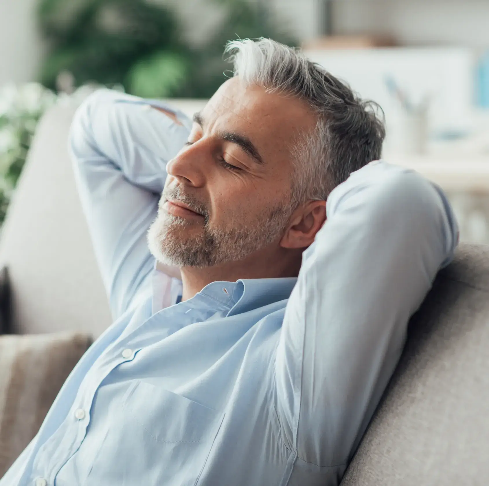Man with gray hair relaxing on a couch, eyes closed, and arms behind his head.
