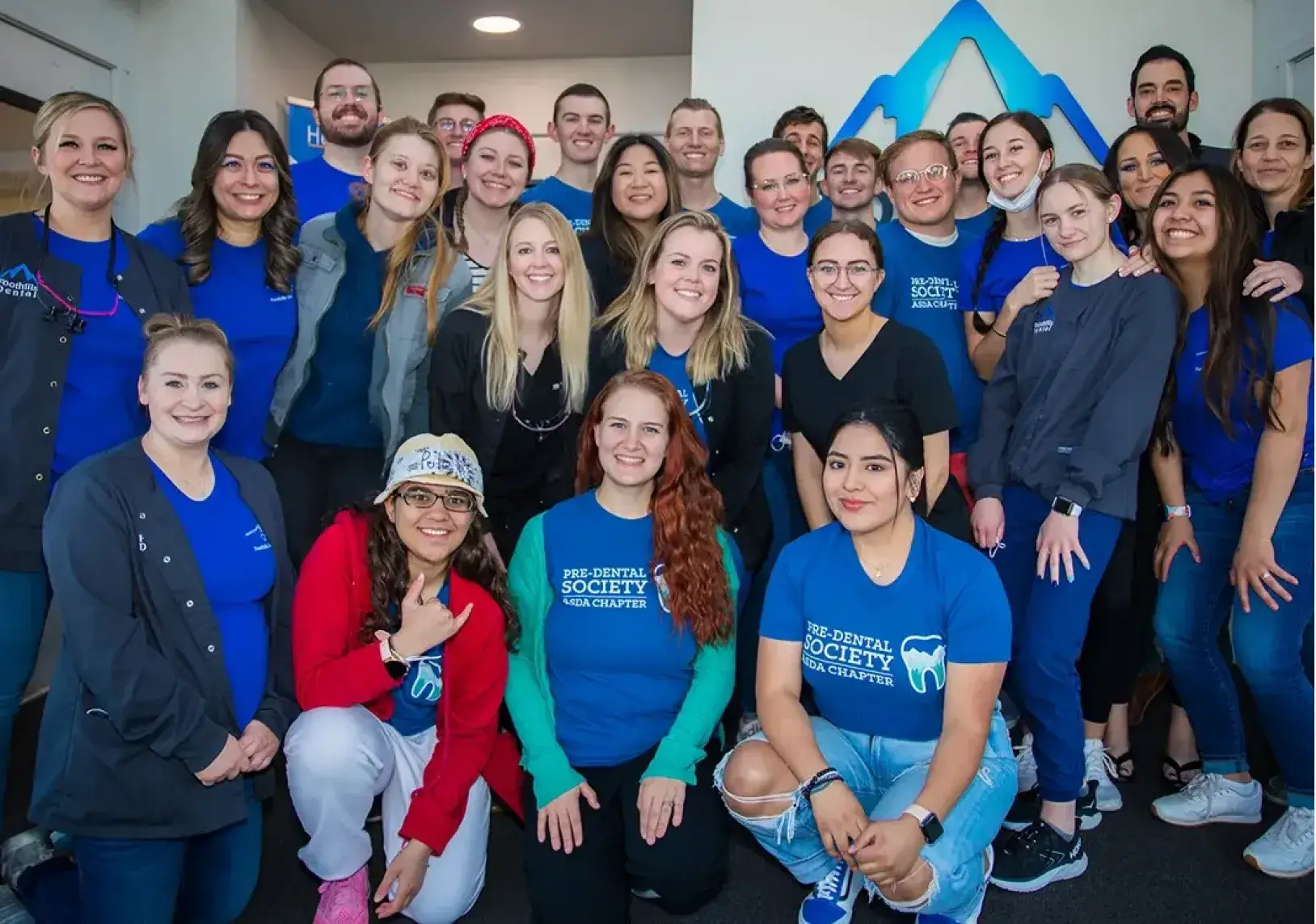 A group of smiling people wearing blue shirts pose together for a group photo indoors.