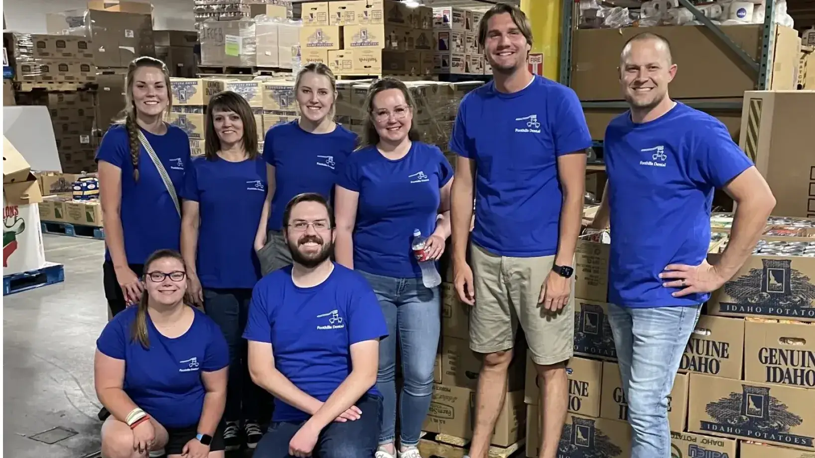 A group of nine people wearing blue shirts pose together in a warehouse with stacked boxes.