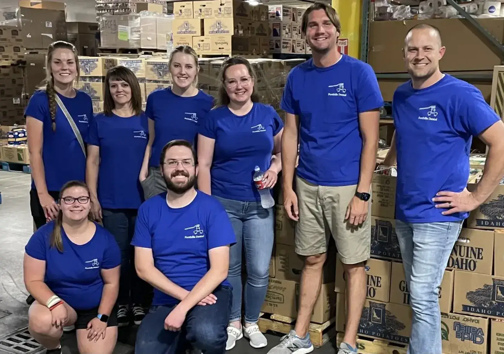 A group of eight people wearing matching blue shirts poses in front of stacked cardboard boxes.