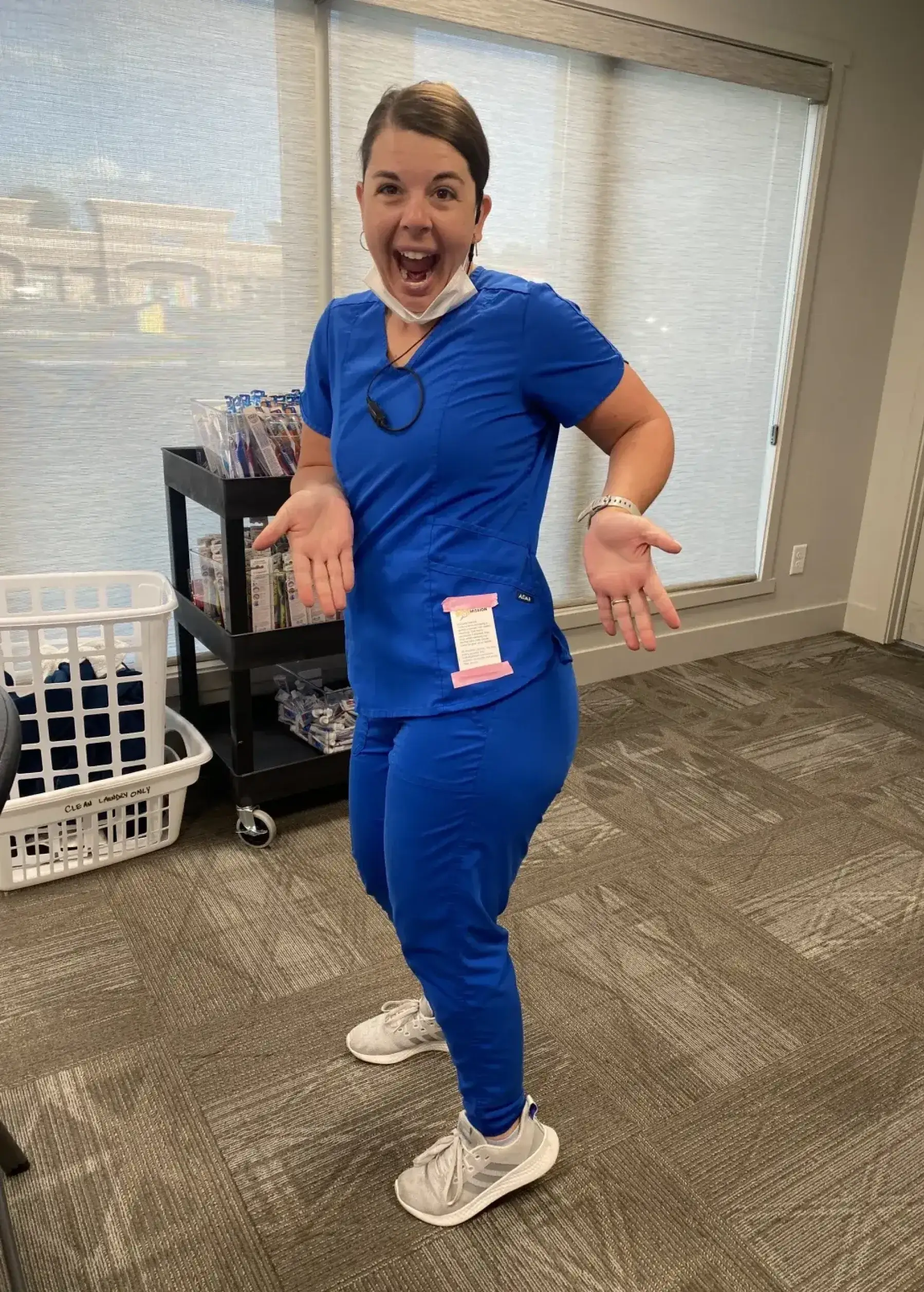 A person in blue scrubs joyfully poses inside an office, with a cart in the background.