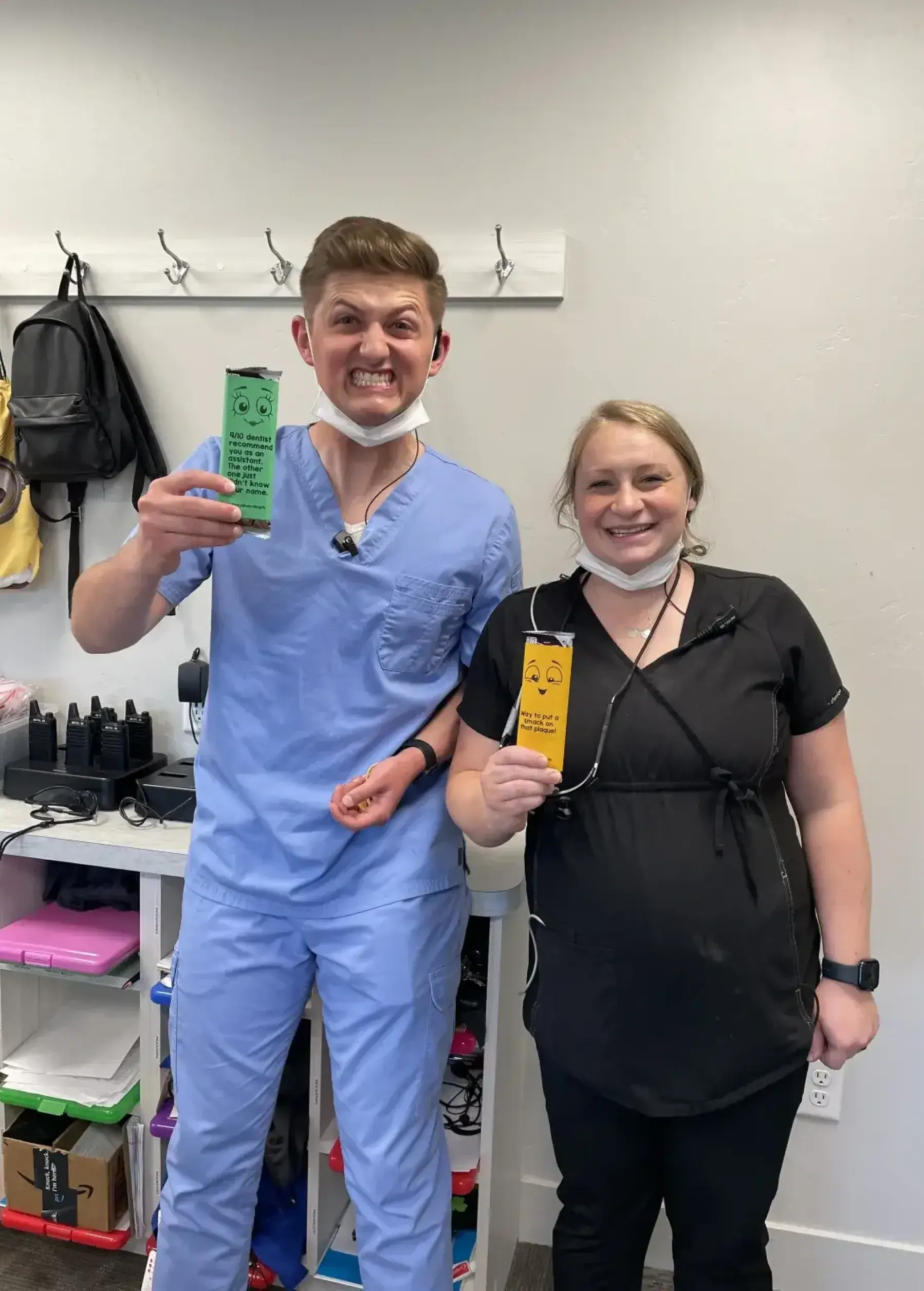 Two smiling dental professionals hold up colorful bookmarks, standing in an office with equipment behind.