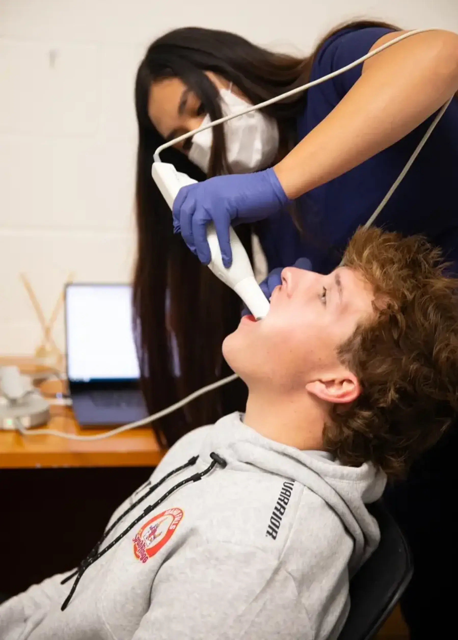 A dental professional uses a handheld tool to examine a seated person's mouth in an office setting.