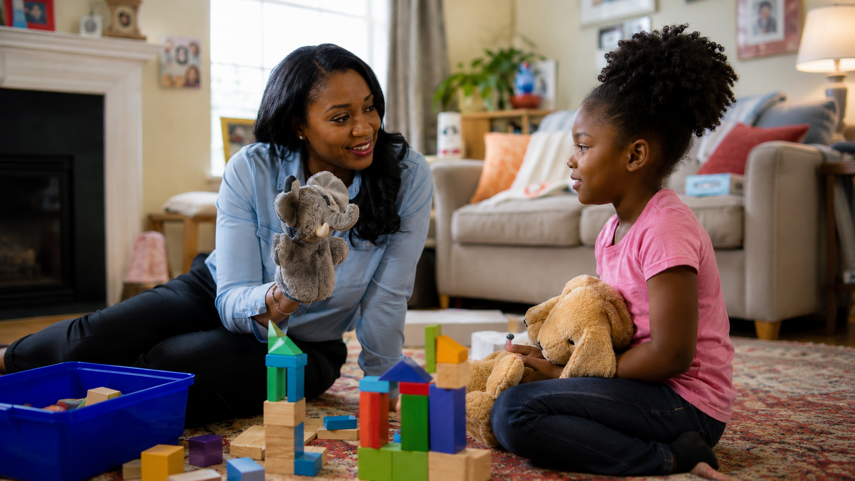 Woman using an elephant puppet to engage a young girl holding a teddy bear while playing with blocks.