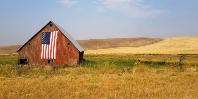 An American flag hangs from a barn that's surrounded by yellowed grass.