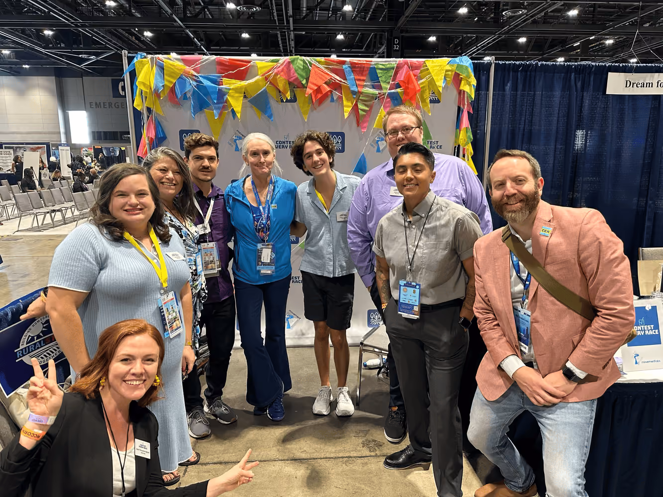 Staff from Contest Every Race wear lanyards and stand in a semi-circle in front of a booth at a conference during the Democratic National Convention.