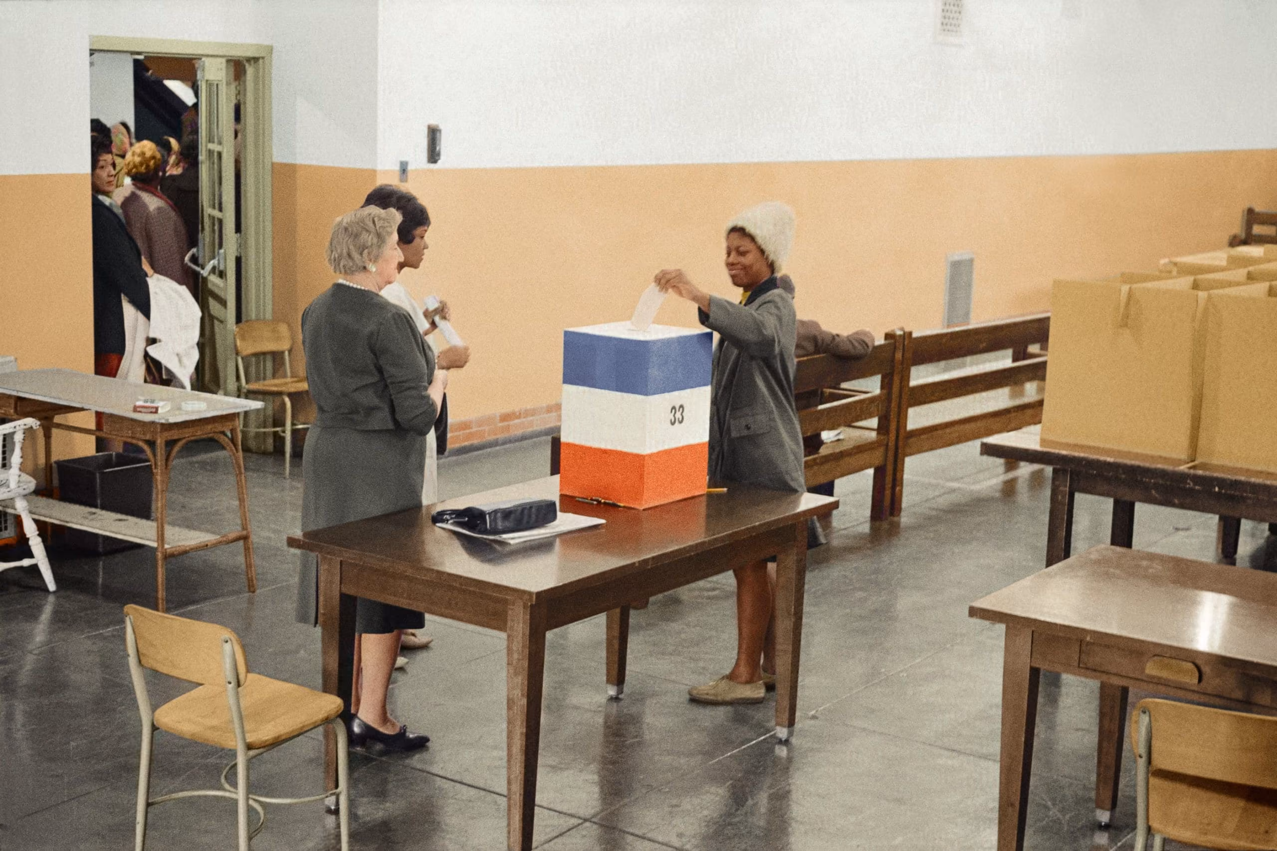 Two women watch another woman place her ballot in a ballot box.
