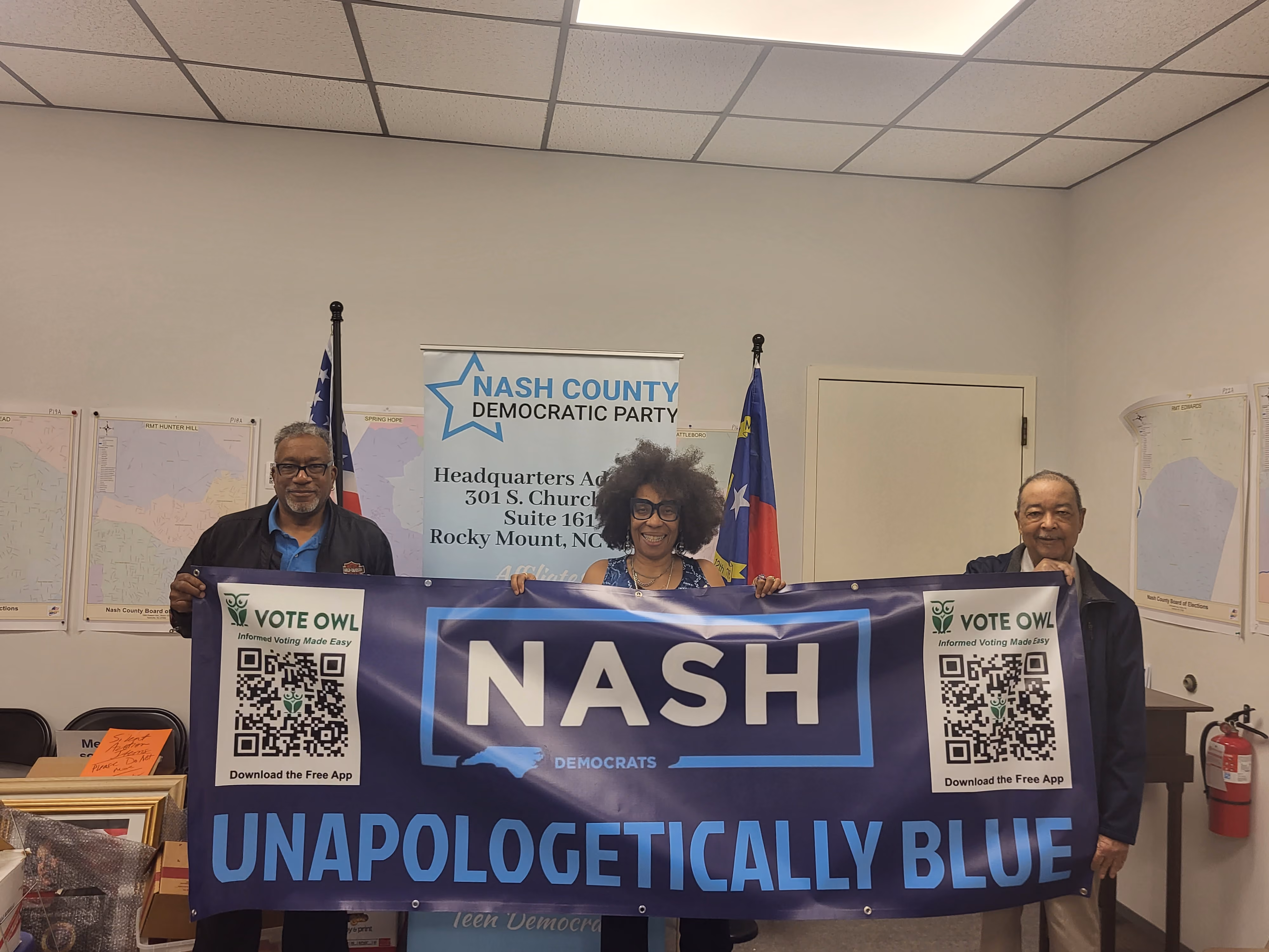 Three members of the Nash County Democratic Party stand in an office holding a banner that says "NASH. UNAPOLOGETICALLY BLUE."
