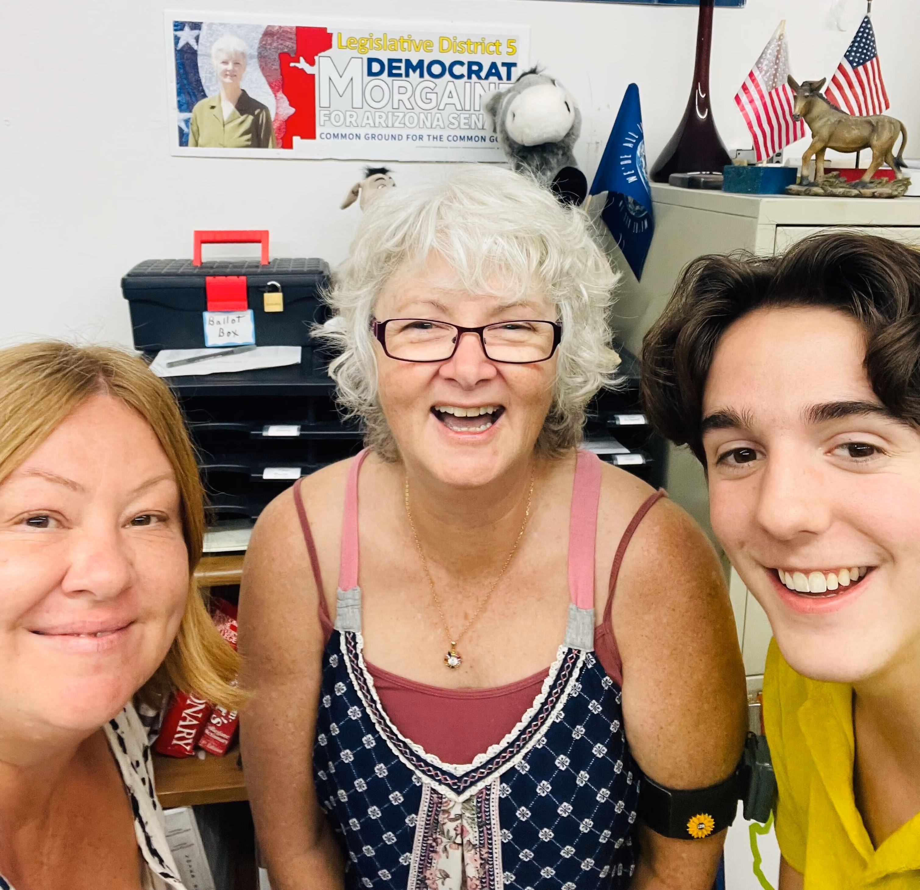 A political candidate smiles to the camera. Beside her are two volunteers, also smiling. Behind them is a sign for that candidate.