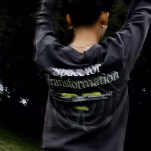 Back view of a person outdoors raising their arms while wearing a dark long-sleeve shirt with the word ‘transformation’ printed across the shoulders.