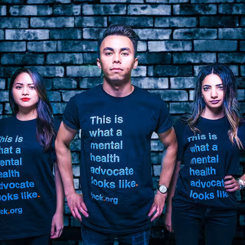 Three young adults stand shoulder to shoulder in matching T-shirts that read ‘This is what a mental health advocate looks like,’ posed against a dark brick wall.