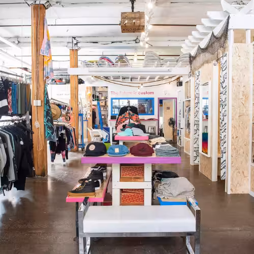 Bright, eclectic retail space with racks of clothing, a central table displaying hats and shoes, and surfboards suspended from the ceiling.