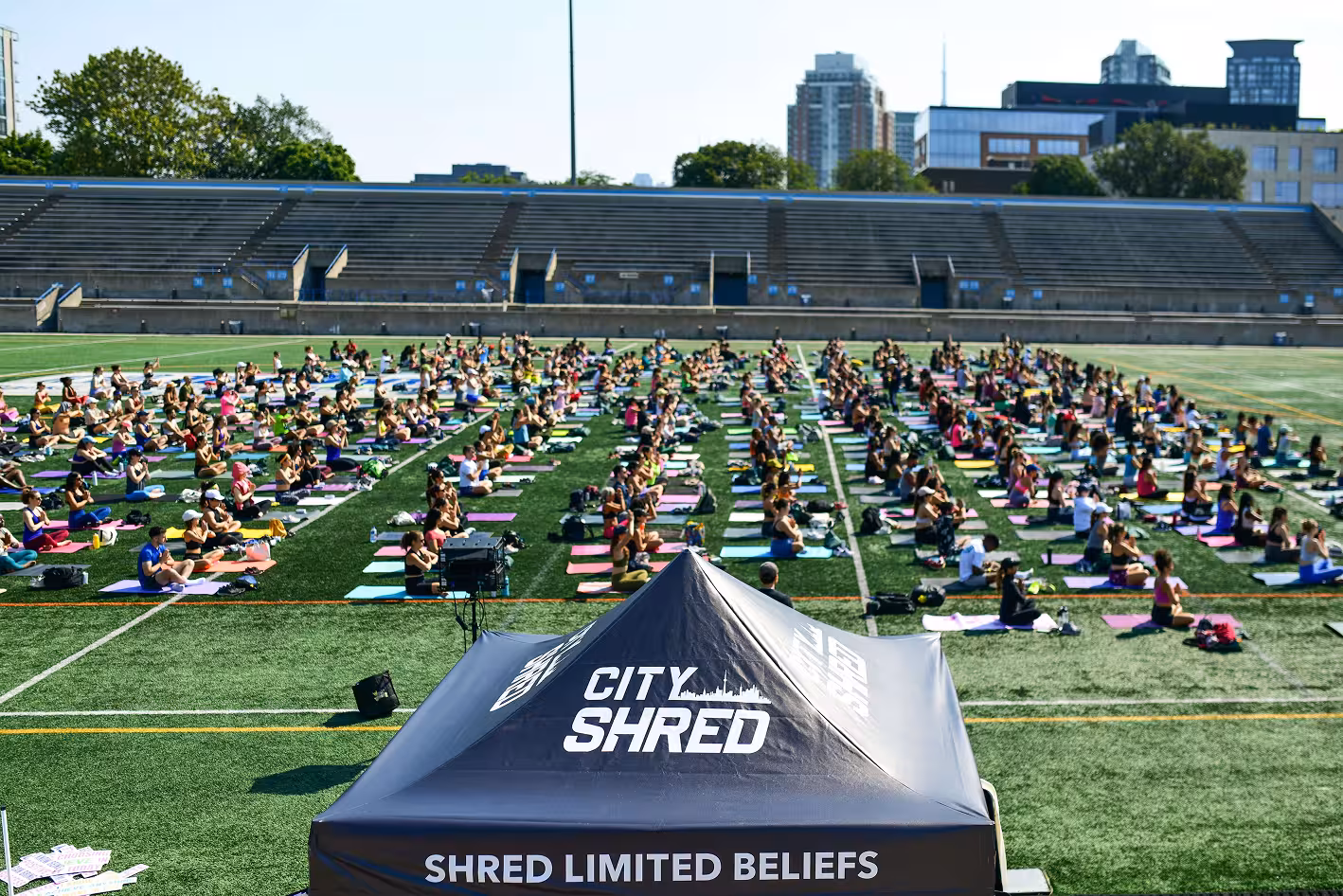 Hundreds of people practicing yoga on mats in a stadium field during a City Shred fitness event, with a tent reading "SHRED LIMITED BELIEFS" in the foreground.