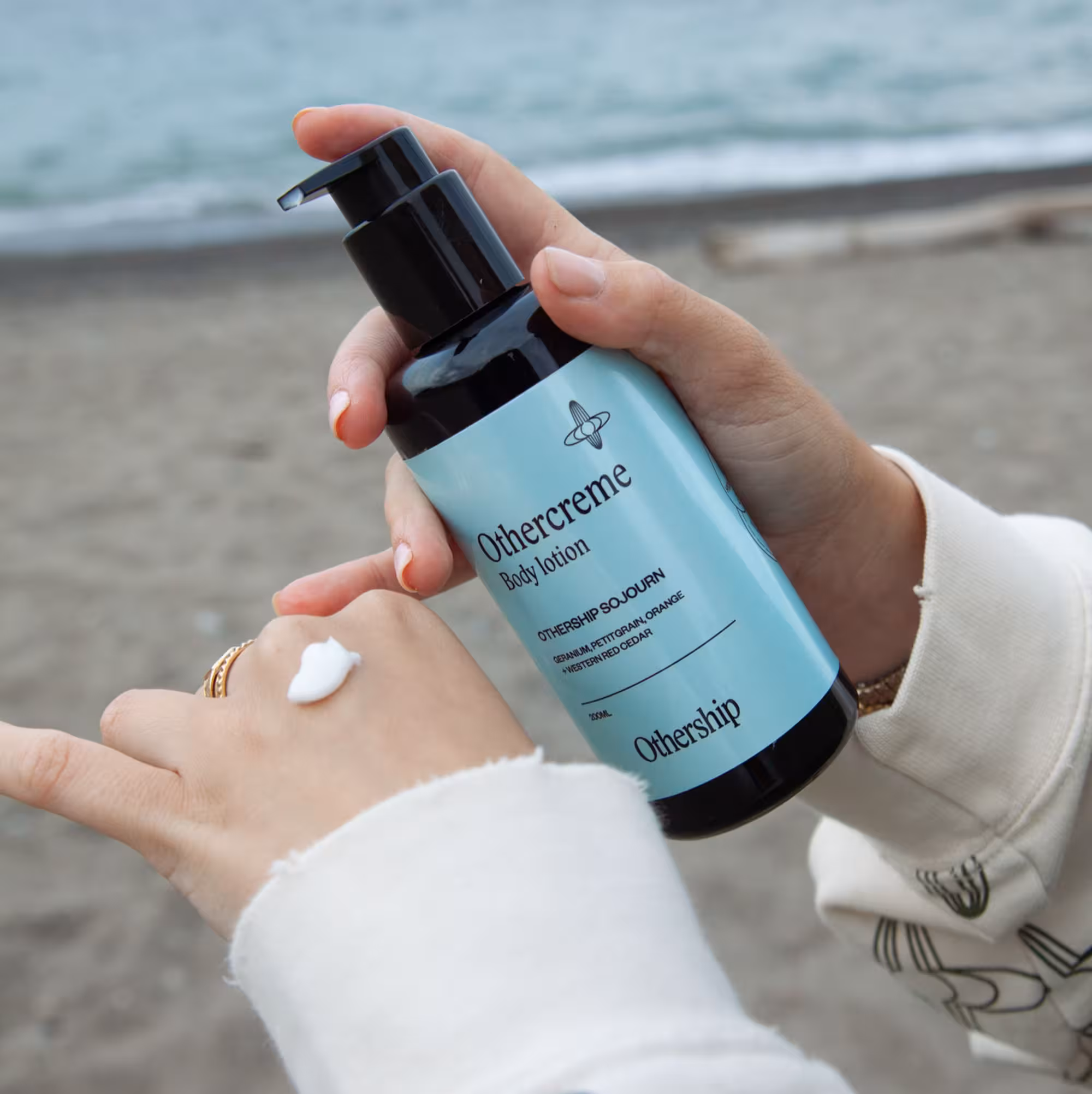 Close-up of hands applying lotion from a blue-labeled bottle of "Othercreme Body Lotion" by Othership, at a sandy beach setting.