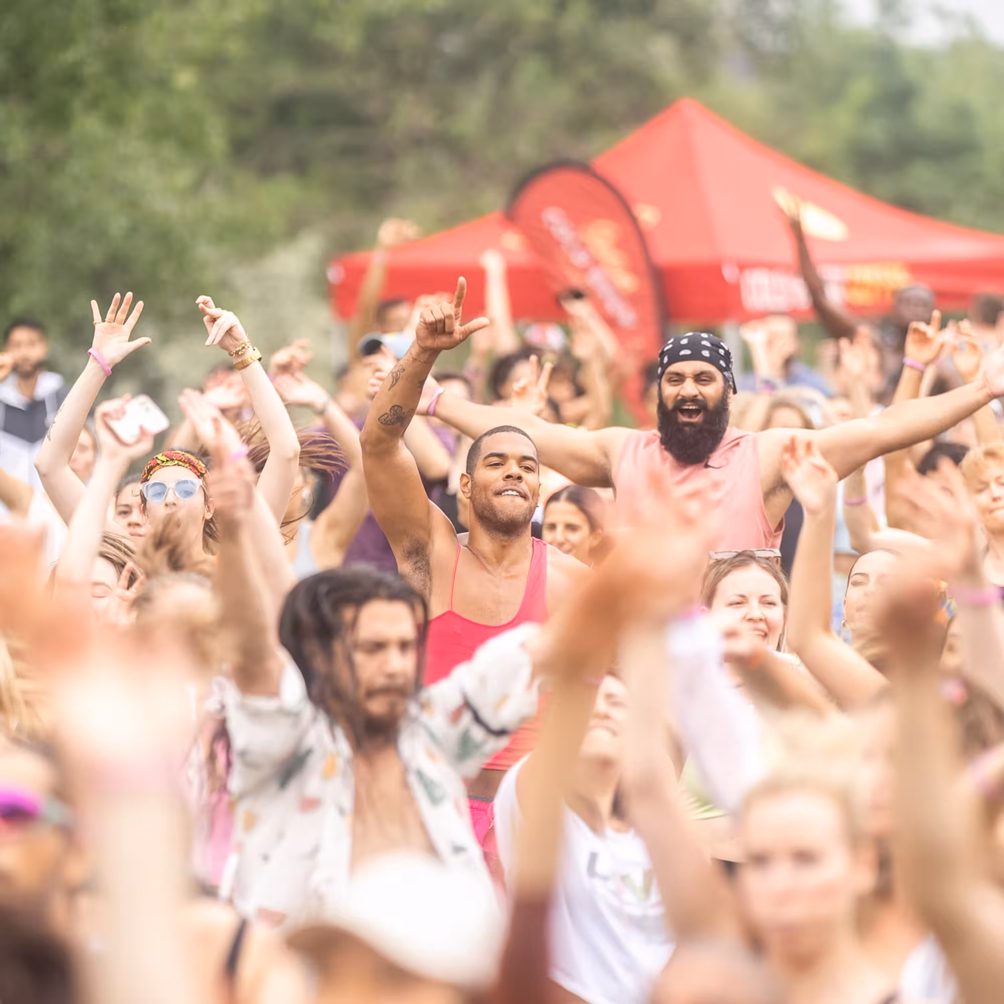 Large joyful crowd at an outdoor event, dancing and raising arms with smiles, colorful outfits, and red vendor tents in the background.