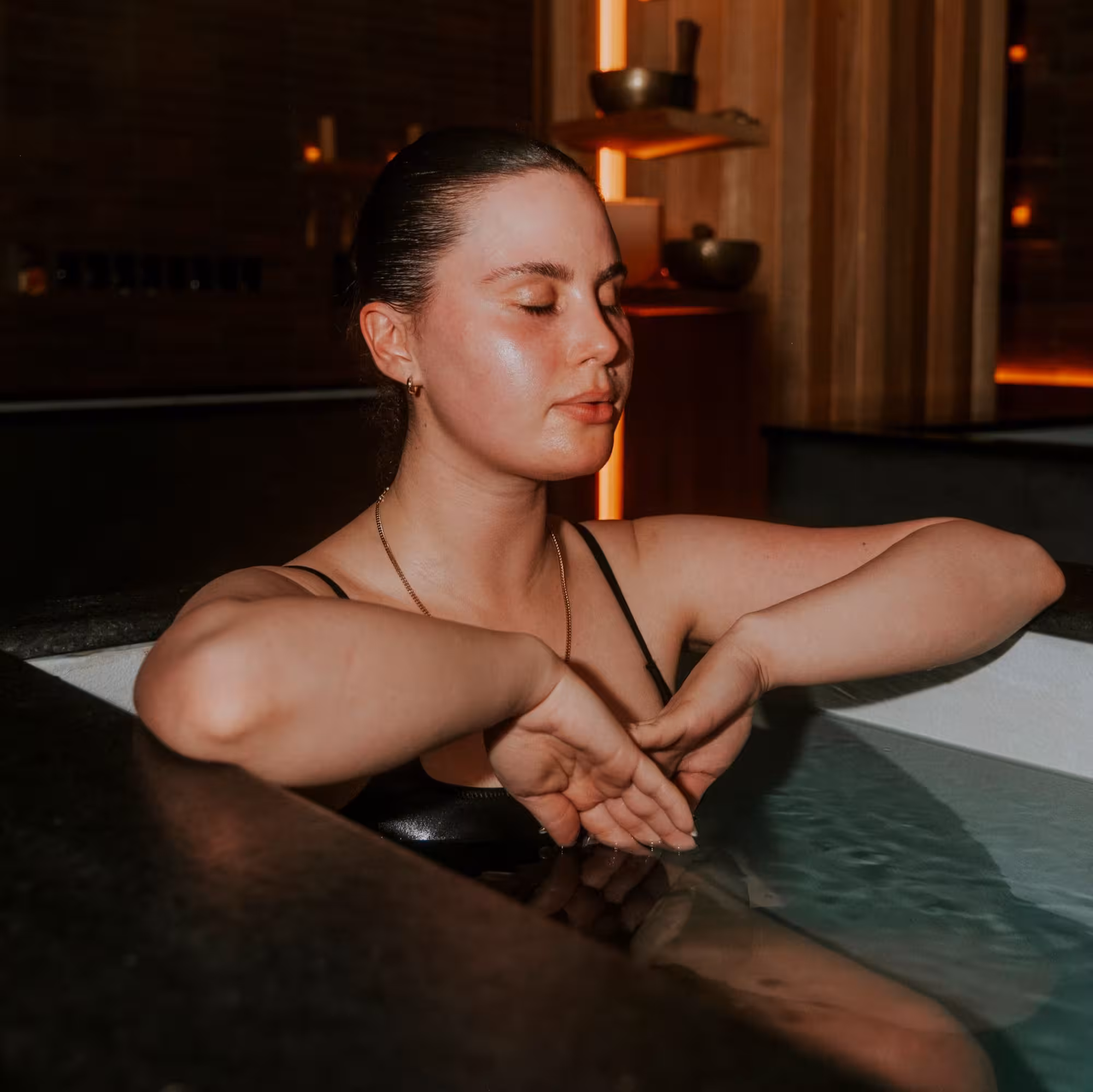 Woman in a black swimsuit with eyes closed, meditating calmly in an indoor ice bath illuminated by warm, ambient lighting.