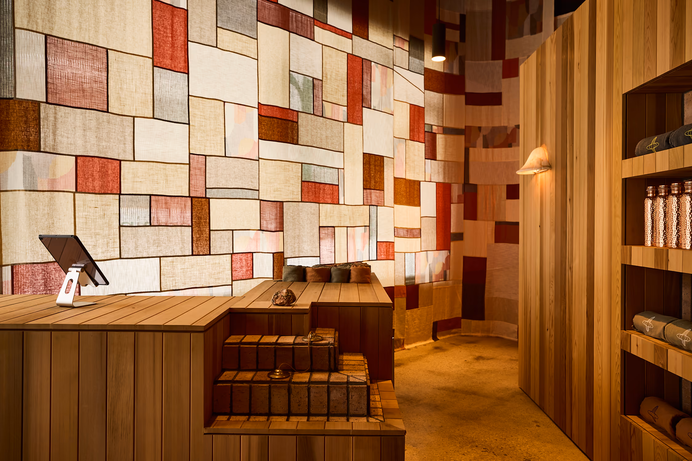 Interior with wooden reception desk, brick steps, colorful patchwork wall art, and wooden shelves with towels and bottles.