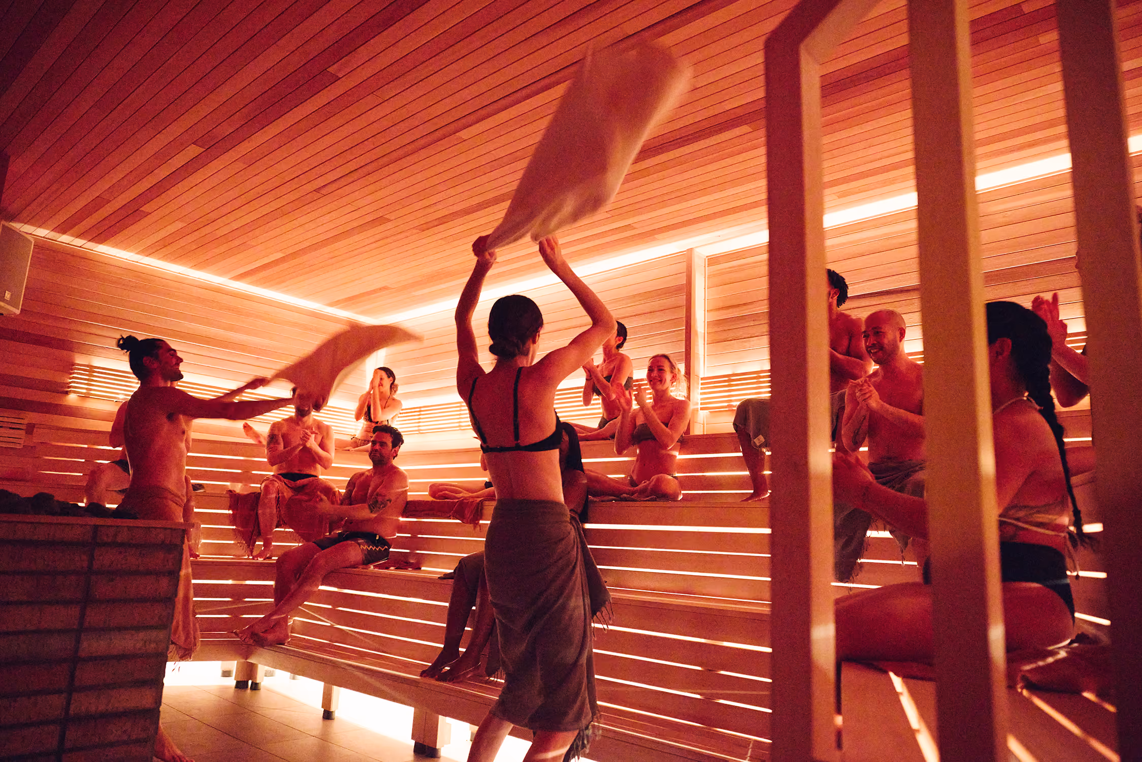 Group of people in swimsuits enjoying a sauna, some waving towels and others clapping on wooden benches.