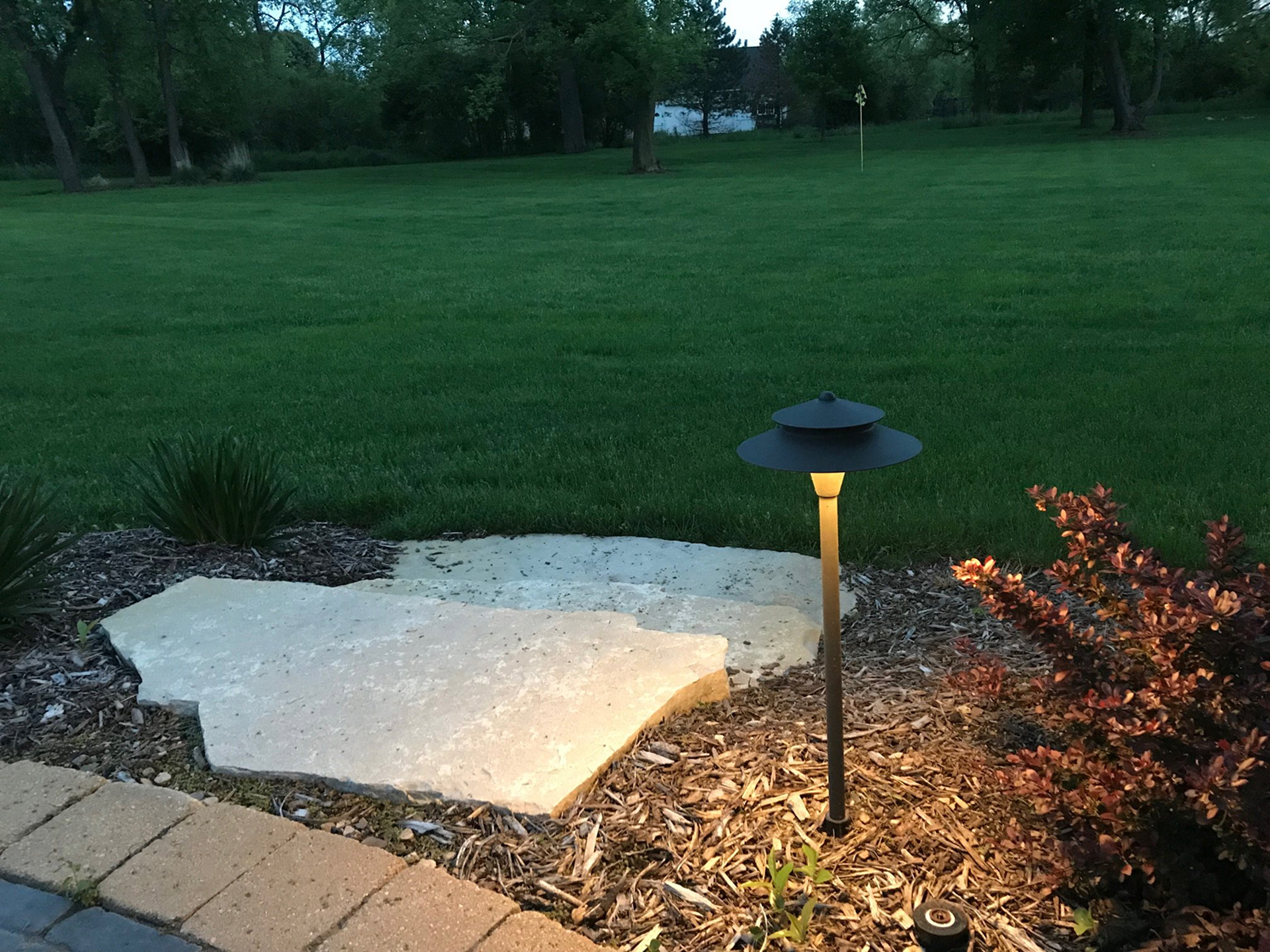 Pathlight softly illuminating a stone paver step surrounded by mulch, ornamental grass, and a landscaped garden bed