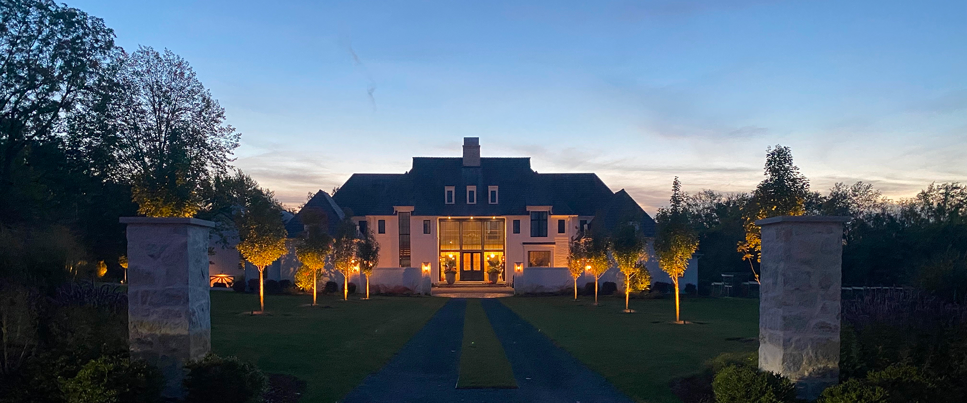 Modern estate with symmetrical uplighting on trees lining the driveway and entryway at dusk