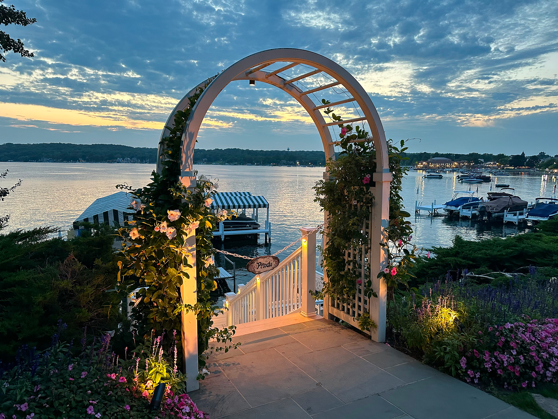 White garden arbor lit with both uplights and downlights at sunset, overlooking a lake dock