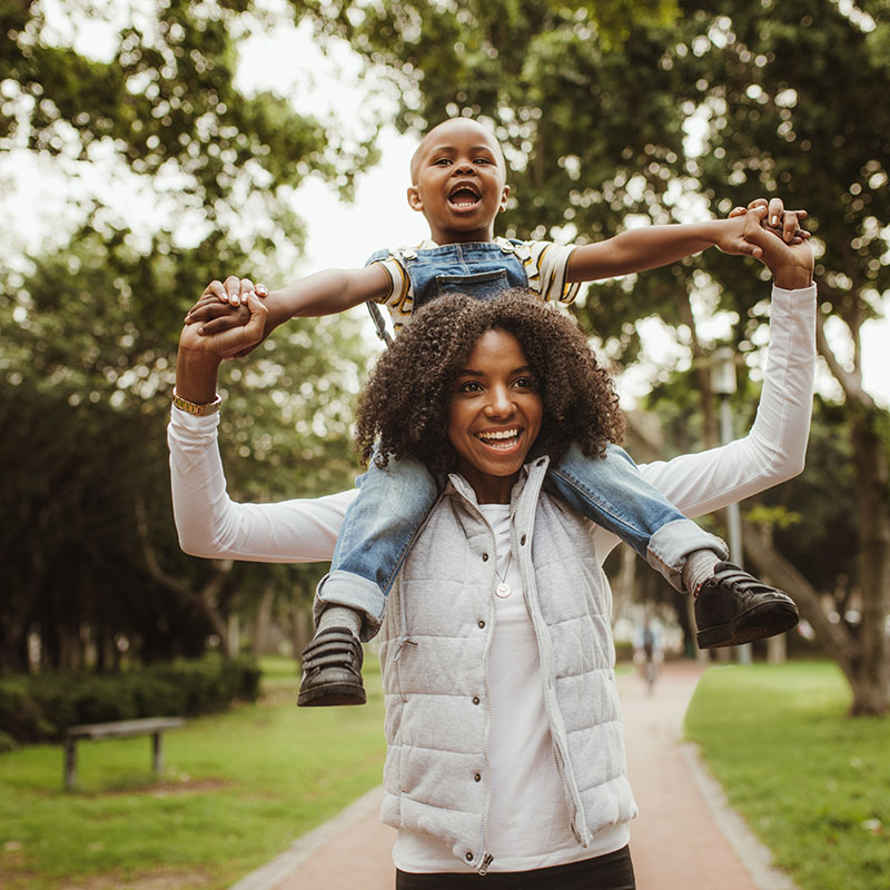 A happy mother walking through a park with her child on her shoulders smiling.