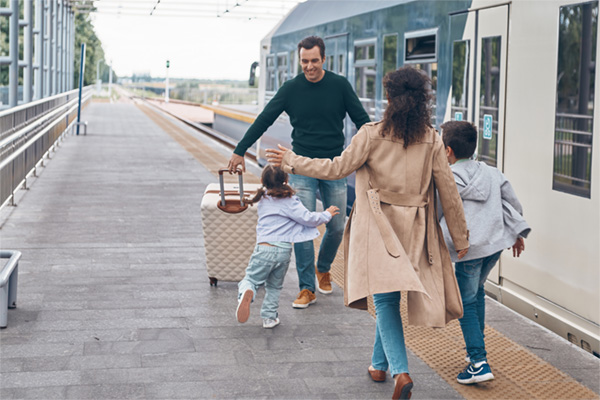 A family reuniting outside at a train station. A man is carrying a suitcase while his two kids and wife run up to him with open arms.
