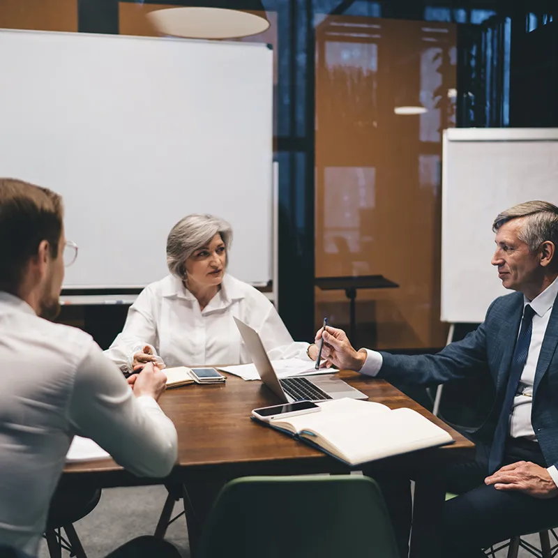 Three business professionals sat at a table in a discussion with whiteboards behind them
